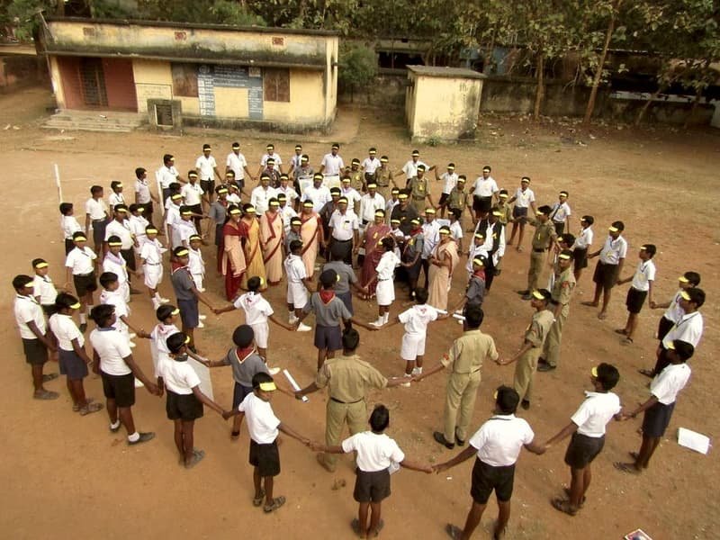 Students and scouts hold hands in a large circle on a school ground during an awareness activity.
